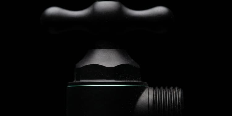 Close-up of a matte black tap with a cross-shaped handle and threaded pipe fitting, lit against a black background.