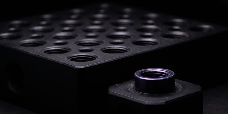 Close-up of a black machined metal plate with a grid of counterbored holes beside a separate hex-shaped fitting with a threaded bore.