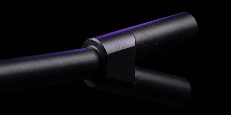 A close-up macro shot of a matte black machined rod splitting into two identical branches at a precision junction; one branch is lit with a thin violet edge of light, the other fades into blurred darkness.