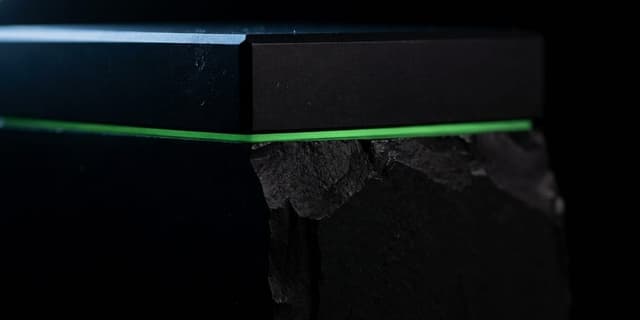 A matte black block split by a single thin green seam, a small finished section above it and a far larger rough mass falling away below