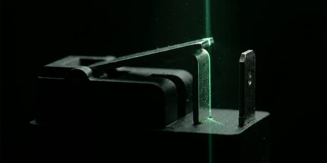 Macro shot of a matte black mechanical relay arm suspended between two contact points on a dark surface, with one contact edge lit by a thin line of green light and the rest of the mechanism fading into black shadow.