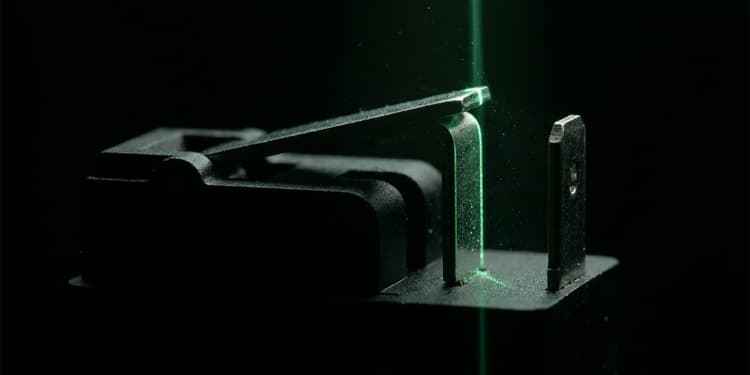 Macro shot of a matte black mechanical relay arm suspended between two contact points on a dark surface, with one contact edge lit by a thin line of green light and the rest of the mechanism fading into black shadow.