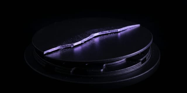 A matte black disc lit from the side by violet light, split by a jagged crack that runs edge to edge, resting on a stack of machined black plates.