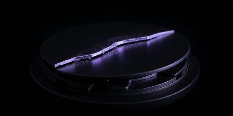 A matte black disc lit from the side by violet light, split by a jagged crack that runs edge to edge, resting on a stack of machined black plates.