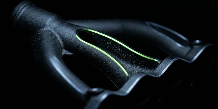 A close-up macro shot of a black machined manifold where one pipe splits into several identical branching channels; only one branch is lit with a thin line of green light along its edge, while the others fade into darkness.