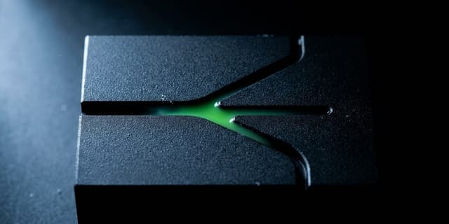 A close-up macro shot of a matte black machined block where a single carved groove splits into several diverging channels of different widths, with a thin line of green light glowing inside the widest branch while the rest of the surface fades into darkness.