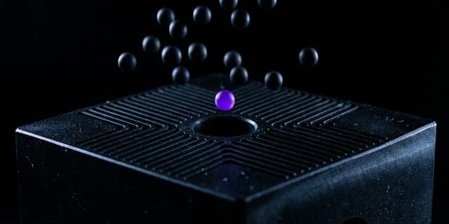 A close-up macro shot of a matte black machined block with many narrow channels converging on one central hole, with numerous small dark spheres falling around it and missing the opening, while a single sphere glowing violet hovers precisely aligned above the hole.