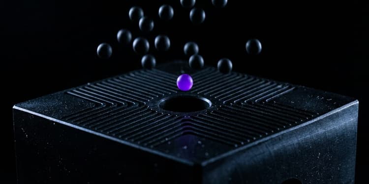 A close-up macro shot of a matte black machined block with many narrow channels converging on one central hole, with numerous small dark spheres falling around it and missing the opening, while a single sphere glowing violet hovers precisely aligned above the hole.