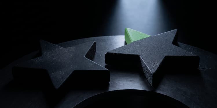 Two matte black star-shaped objects on a dark surface, with one point of the rear star painted green, lit by a soft beam from above against a black background.