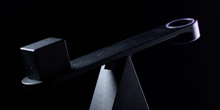 A black seesaw-like balance beam with a cube on one end and a circular ring rimmed in purple light on the other, resting on a triangular fulcrum against a black background.