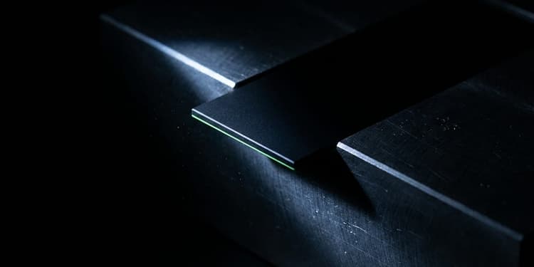 A thin black panel with a glowing green edge emerges from a dark machined slot and hangs suspended mid-drop over empty darkness, with no surface beneath to catch it.