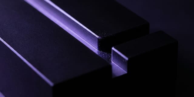 A close-up of a matte black machined block with a single groove splitting into two channels; one channel glows violet along its length, while the other channel remains dark and ends abruptly at a sharp edge.