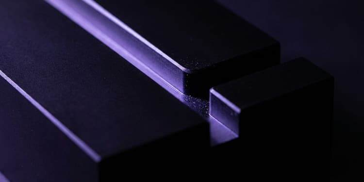A close-up of a matte black machined block with a single groove splitting into two channels; one channel glows violet along its length, while the other channel remains dark and ends abruptly at a sharp edge.