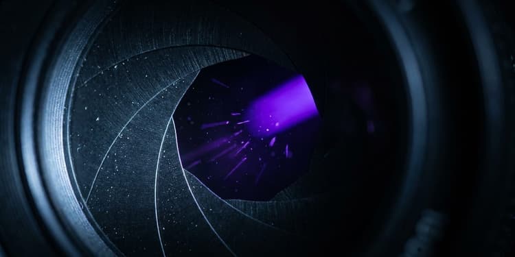 Close-up macro photograph of a black mechanical iris aperture with thin blades opening, a narrow beam of violet light splitting into several thinner violet slivers as it spills through the widening gap, set against a completely black background.