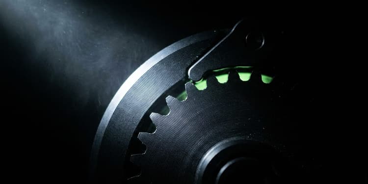 Extreme close-up of a black machined gear and ratchet mechanism in near-total darkness, with the pawl lifted away from the gear teeth so it spins without engaging; a thin sliver of green light glows in the gap between pawl and teeth, and the rest of the frame fades to black.