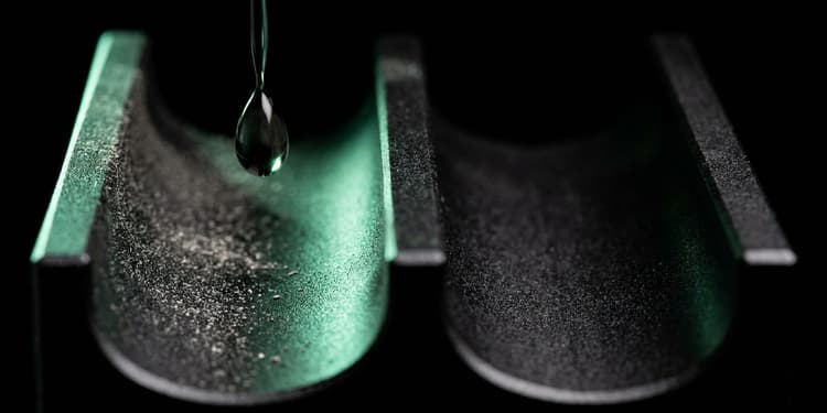 A close-up macro shot of a matte black machined surface splitting into two narrow channels; one channel holds a single green-lit droplet suspended mid-fall, while the other channel is empty and fades into darkness.